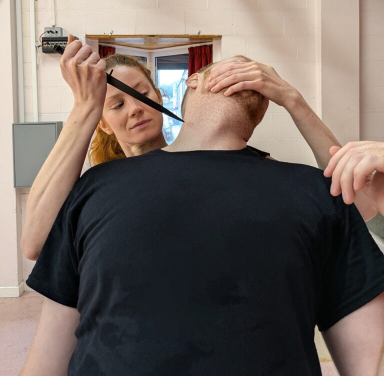 Gisela, a female member of Battalion Martial Arts, executing a sentry takedown on another student. She has sneaked up behind the student, gripping their mouth to turn their head aside, while holding a dagger to their throat