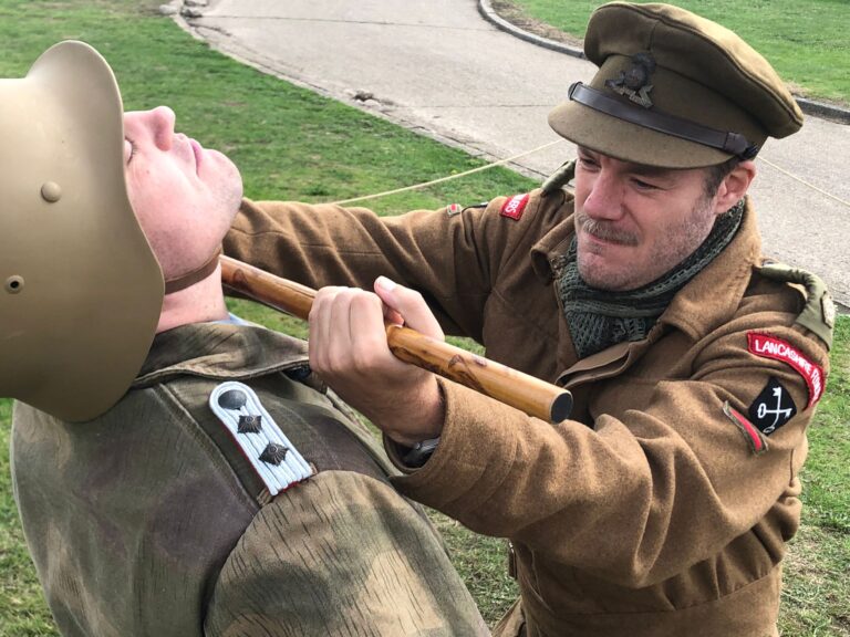 A man in British WW2 uniform attacks a Man in German WW2 Uniform by slamming a stick into his throat as part of a Reenactment event