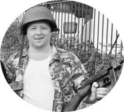 A Picture of an instructor of Battalion Martial Arts Mike portraying a US Sea Bee. He is smiling at the camera dressed in a Hawaiian shirt and US military helmet holding a Thompson sub machine gun.